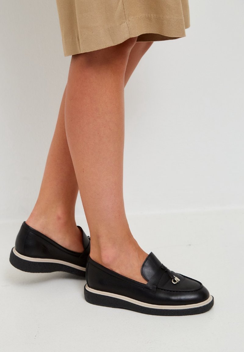 Femme portant des mocassins en cuir noir avec un détail de boucle argentée, assortis à une jupe beige, debout sur un sol blanc.