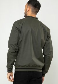 Homme vu de dos portant une veste bomber vert foncé avec des bordures noires et un pantalon noir, sur un fond clair uni.