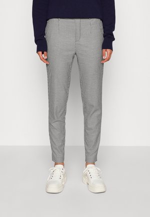 Person iført sort-hvide ternede slim-fit bukser, hvide chunky sneakers og en mørk langærmet top, stående mod en ensfarvet baggrund.