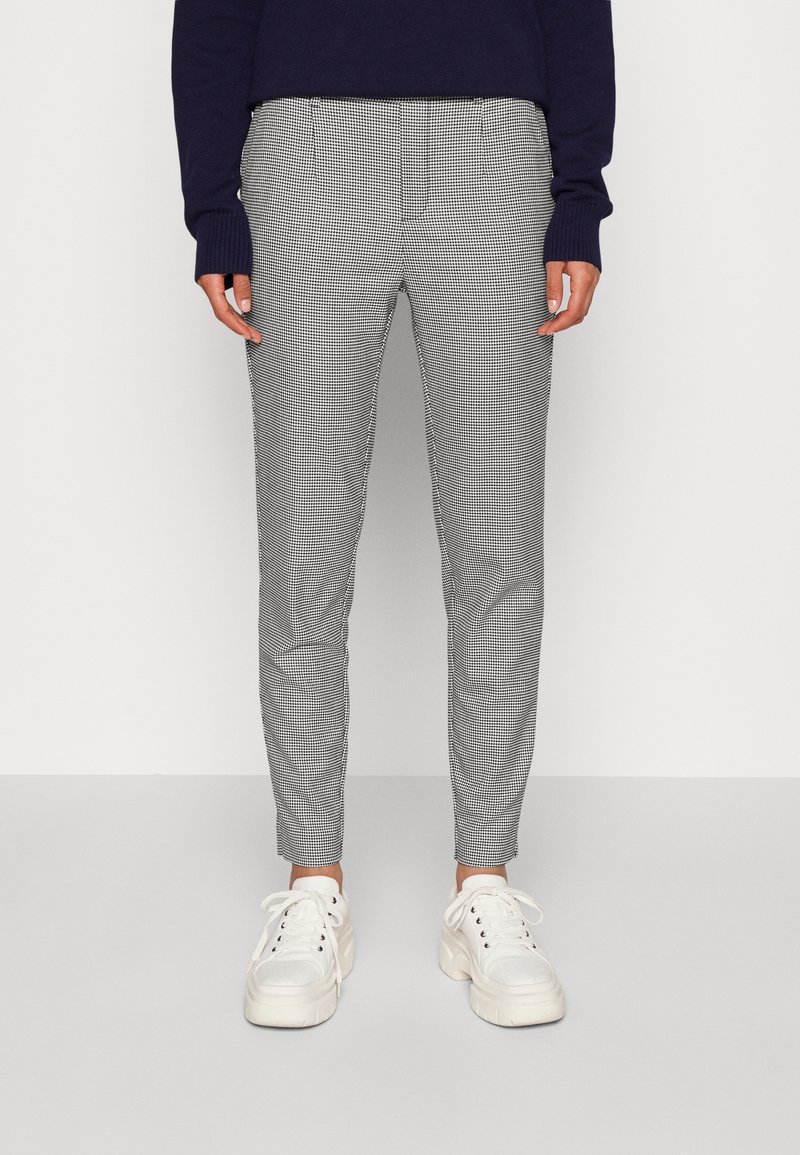 Personne portant un pantalon slim à carreaux noir et blanc, des baskets blanches épaisses et un haut à manches longues sombre, debout devant un fond uni.