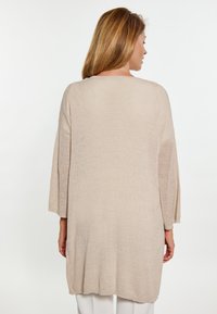 Beige stickad cardigans med långa ärmar, lös passform och texturerad tyg. Bakdelen har en subtil lyster och en böjd nederkant.