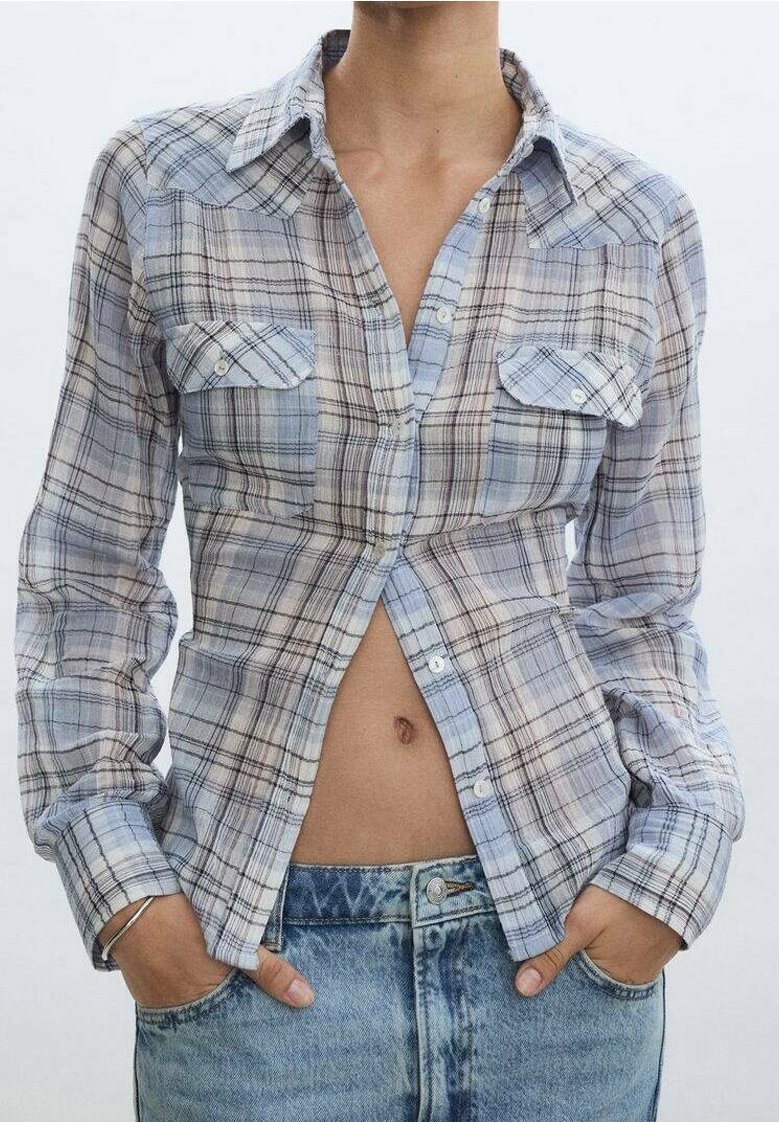 Personne portant une chemise à carreaux bleue et grise boutonnée, ouverte en bas, montrant le nombril, les mains dans les poches d'un jean en denim bleu clair.