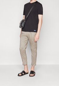 Man in zwarte t-shirt, beige cropped broek, zwarte slide-sandalen en zwarte schoudertas tegen een effen achtergrond.