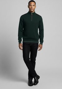 Jersey de punto acanalado verde oscuro con cuello de cremallera de cuarto, combinado con vaqueros ajustados oscuros y botines negros.