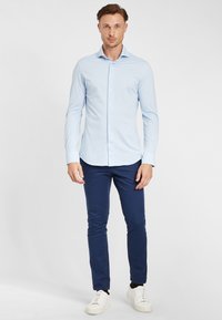 Camicia azzurra a maniche lunghe con colletto a bottoni, abbinata a pantaloni slim-fit blu navy scuro e sneakers bianche. Tessuto dalla texture liscia.
