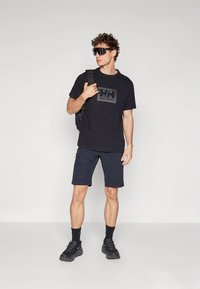 Juodas marškinėliai su stačiakampiu logotipu, tamsiai mėlynos trumpos kelnės, juodi sportbačiai ir akiniai nuo saulės. Asmuo laiko kuprinę; minimalistiškas dizainas ir spalvos.