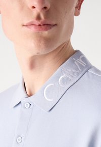 Polo bleu clair en tissu texturé, avec un col et un logo brodé "Calvin" en blanc sur le côté gauche. Deux boutons visibles.