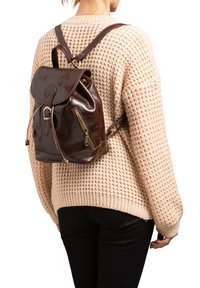 Bruine leren rugzak met een trekkoordsluiting, een decoratieve gesp en zijvak met ritsen, gedragen boven een crèmekleurige gebreide trui.