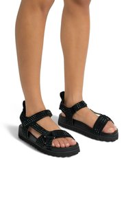 Sandalias negras con acentos de tachuelas, que cuentan con múltiples correas ajustables, una suela de goma gruesa y un diseño de punta abierta.