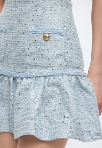 Φούστα από ανοιχτό μπλε τουίντ με χρυσές και ασημένιες παγιέτες, διακοσμητική τσέπη με κουμπιά και βολάν στο κάτω μέρος, σε χαμηλό μέρος σώματος ατόμου.