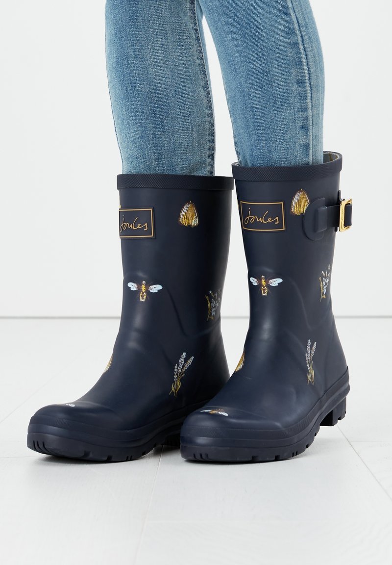 Bottes de pluie bleu marine avec des motifs d'abeilles, de coquillages et floraux, portées avec un jean bleu clair sur un sol blanc.