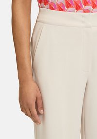 Ljusbeige byxor med en slät textur, prydda med framveck och snedställda fickor, kompletterade av en färgglad blommönstrad topp.