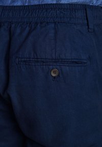 Pantaloni corti blu navy con vita elastica e una tasca posteriore con chiusura a bottone marrone. Il materiale sembra leggero e liscio.