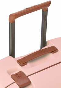 Valigia rigida rosa con maniglia testurizzata marrone e barra di estrazione. Presenta un pulsante per assicurare la chiusura. Design moderno.
