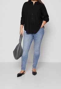 Schwarze Hemdjacke, blaue Skinny-Jeans, graue Handtasche und schwarze flache Schuhe mit spitzem Zehen und akzentuierender Hardware. Glatte Stofftexturen.