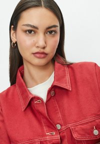 Red denim jacket with a collar, two chest pockets, silver-tone buttons, and contrasting white stitching, paired with a white shirt underneath.