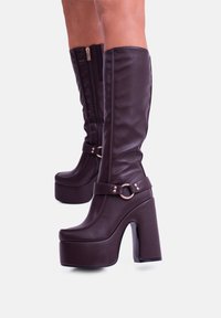 Stivali alti fino al ginocchio in faux pelle marrone con una piattaforma, tacco spesso, zip laterale e dettaglio decorativo a fibbia sulla cinghia alla caviglia.