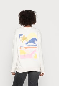 Sudadera blanca con un estampado colorido en la espalda, que muestra una palmera, olas y formas abstractas en amarillo, azul, rosa y naranja.