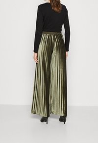 Kalhoty s vysokým pasem a záhyby v olivově zeleném sametu, s širokým střihem nohavic, doplněné přiléhavým černým topem a vysokými podpatky.