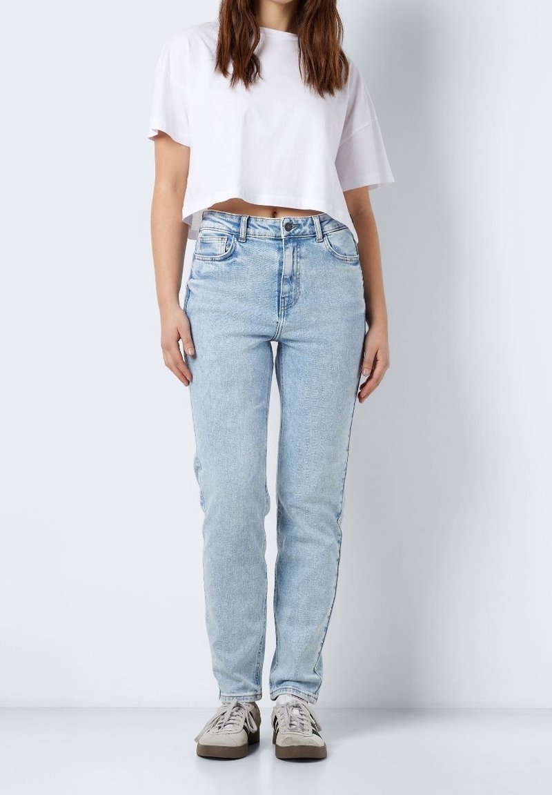 Bílá zkrácená trička s krátkým rukávem, kombinovaná se světle modrými džíny rovného střihu. Obuv má šedé a béžové akcenty.