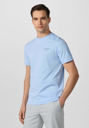 Junger Mann trägt ein hellblaues H.Sport T-Shirt und hellgraue Hose, steht mit einer Hand in der Tasche vor einem einfarbigen Hintergrund.