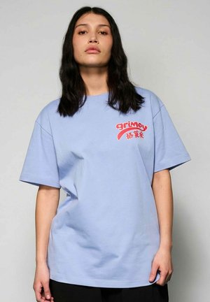 Frau mit langen dunklen Haaren, die ein hellblaues, übergroßes T-Shirt mit rotem "grimey" Schriftzug und asiatischen Schriftzeichen auf der Brust trägt, stehend vor einem schlichten Hintergrund.