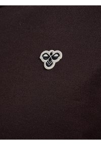 Sort stof med en struktureret overflade, der har et lille broderet bi-logo i hvidt og sort placeret på venstre side.