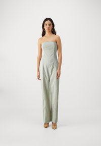 Combinaison sans bretelles en tissu vert clair, avec jambes droites, corsage ajusté et détails de couture minimalistes. Portée avec des chaussures beiges.