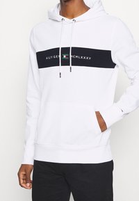 Witte hoodie met een zwarte horizontale streep met tekst en een logo in groen, gemaakt van zacht stof met een voorkantzak en verstelbare trekkoorden.