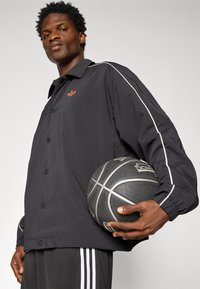Veste de sport noire avec un logo rouge, dotée de rayures blanches sur les côtés. Le mannequin tient un ballon de basket noir et porte un pantalon noir avec des rayures blanches.