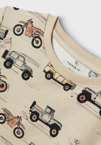 Beige huppari, jossa on eri ajoneuvojen printti, mukaan lukien rekkoja ja moottoripyöriä, yksityiskohtaisilla yksityiskohdilla ja ribatulla kaula-aukolla.