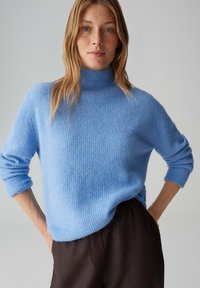 Pull à col roulé bleu clair côtelé, fabriqué en matière douce et texturée. Caractérisé par des manches longues et une coupe décontractée, associé à un pantalon sombre.