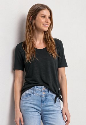 Jeune femme aux longs cheveux châtain clair portant un haut noir à manches courtes noué à la taille et un jean bleu clair, souriante et regardant vers la droite.