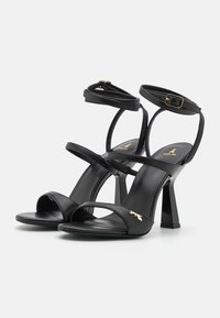 Sandales noires à talons hauts avec de fines lanières à la cheville et un design à bout ouvert. Présente un accent doré et un talon carré et robuste. Texture en cuir lisse.