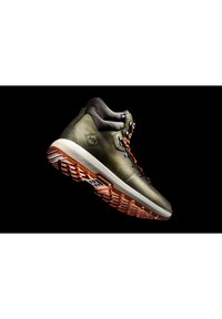 Bota de senderismo verde con una superficie lisa, suela de goma marrón y cordones multicolores. Presenta acentos metálicos y un diseño de soporte en el tobillo.