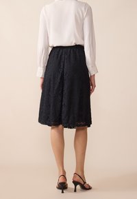 Hame lace hitam dengan pola bunga, sepanjang lutut dengan tepi scalloped. Dipadukan dengan blus lengan panjang putih dan sandal hak hitam.