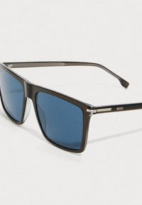 Ορθογώνια μαύρη-περικεφαλίδα γυαλιά ηλίου με μπλε φακούς και ασημένιες λεπτομέρειες στους βραχίονες, με την επιγραφή "BOSS" στο πλάι.