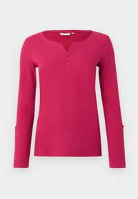 Langærmet lyserød bluse med ribbet tekstur, med en Henley-udskæring og tre knapstolper. Inkluderer slidsdetaljer ved ærmernes kanter.
