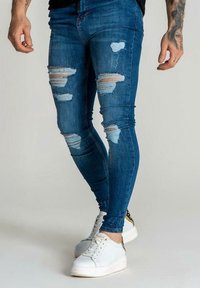 Kapeet siniset farkut, joissa on kuluneita yksityiskohtia, useita repeämiä ja vaaleansinisiä lapuja, yhdistettynä valkoisiin tennareihin.