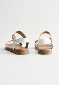 Sandales blanches avec une finition lisse, dotées de deux brides à l'avant et d'une bride de cheville avec une boucle en or, et une semelle marron.