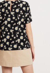 Svart blommig blus med vita prästkragar och gula mittpunkter; korta ärmar, rundad nederkant och detaljer med nyckelhål på baksidan. Matchad med en beige kjol.