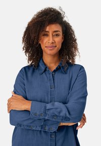 Donna con capelli ricci che indossa una camicia azzurra a bottoni, in piedi con le braccia incrociate e un leggero sorriso su uno sfondo bianco uniforme.