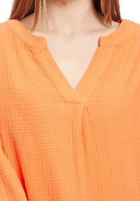 Top a V scollatura di un vivace arancione, realizzato in tessuto testurizzato. Presenta una vestibilità rilassata con sottili dettagli delle cuciture lungo il collo e il corpo.