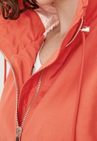 Orange Zip-Jacke mit Kapuze, ausgestattet mit einem silbernen Reißverschluss, weichem Material und leichter Innenauskleidung. Nahaufnahme der Reißverschluss- und Kragen Details.