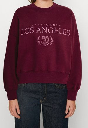 Bordowa bluza z napisem „California Los Angeles” i emblematem, noszona z ciemnoniebieskimi jeansami przez stojącą osobę widoczną od szyi do bioder.