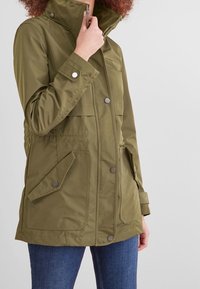 Giacca impermeabile verde oliva con colletto alto, due tasche frontali e chiusure a scatto; presenta una vita cintrata e una texture liscia.