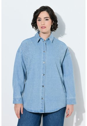 Giovane donna con capelli corti e scuri che indossa una camicia di jeans azzurro chiaro con borchie argentate nella parte superiore davanti e jeans blu scuro.