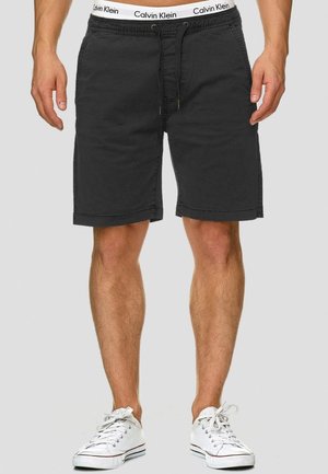 Man draagt zwarte knielange shorts met trekkoord, witte Calvin Klein tailleband zichtbaar, en witte sneakers tegen een effen achtergrond.