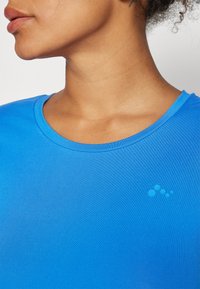 Blå atletisk topp med en slät textur, rund halsringning och korta ärmar. Har en liten, ljusblå logotypdetalj på bröstet.