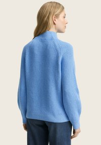 Pull en maille côtelée bleu avec un col haut, présentant une coupe décontractée et des manches longues. Texture douce, montré de dos, associé à un jean.